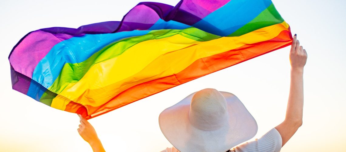 Back view of woman in white dress and hat holding gay pride flag at sunset in beach