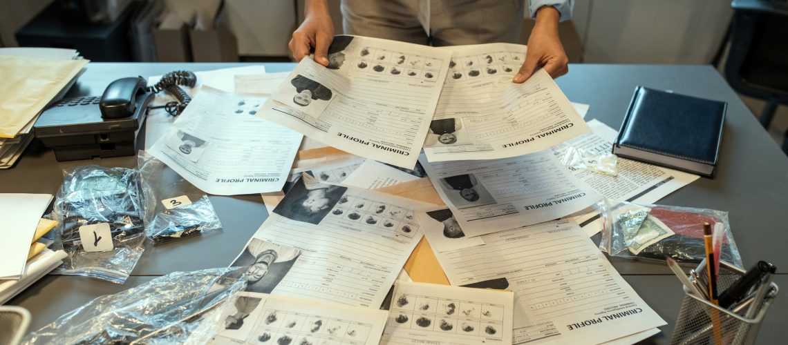 Hands of female investigator holding criminal profiles over desk with documents